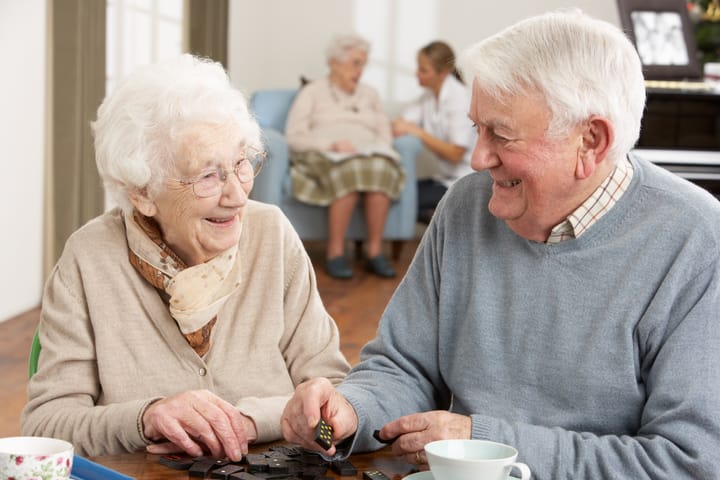 Two older people smiling and looking at each other playing a game of dominoes. In the background another older lady is being spoken to by a care worker