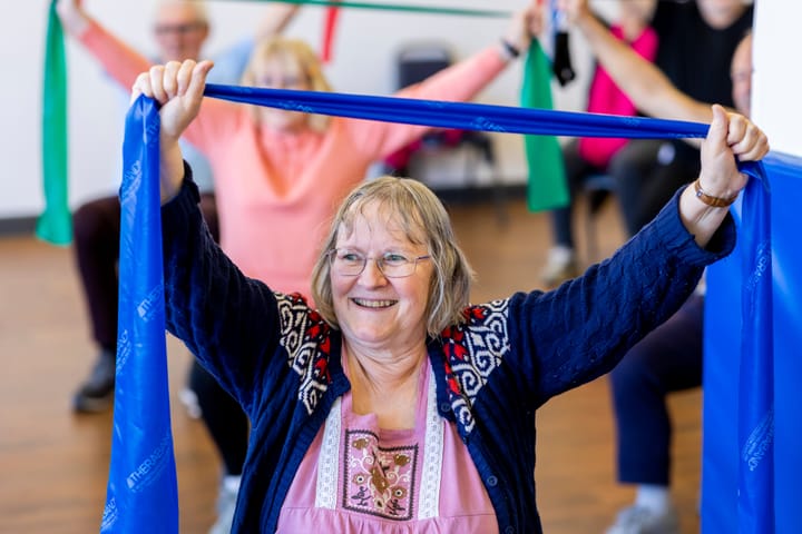 An older lady in an exercise class with a blue stretch band over her head. In the background other older people are doing then same exercise