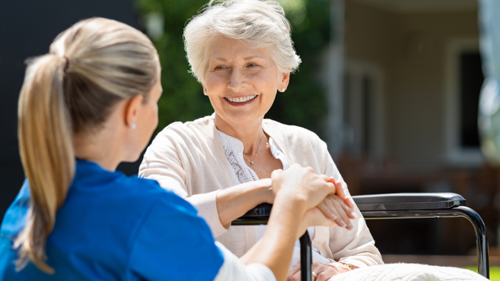Women sat in a wheelchair in her garden smiling at a carer who is crouched down talking to her.