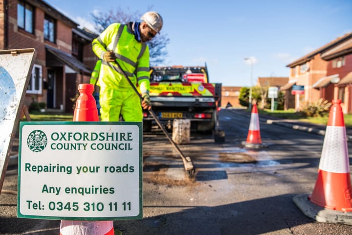 Highways worker in safety gear brushing over a repaired pothole in a residential street. He is surrounded by cones with a repairing your roads sign in the foreground with the enquiries number